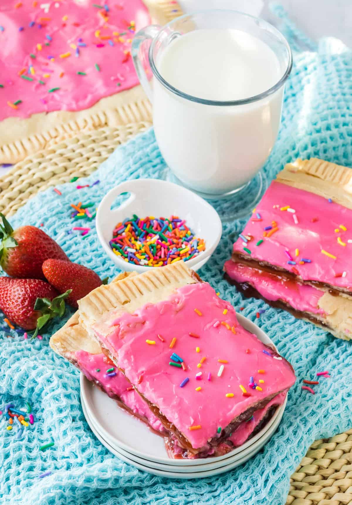 Pieces of a homemade sheet pan pop tart on a plate with more pieces, glass of milk, and sprinkles in the background.