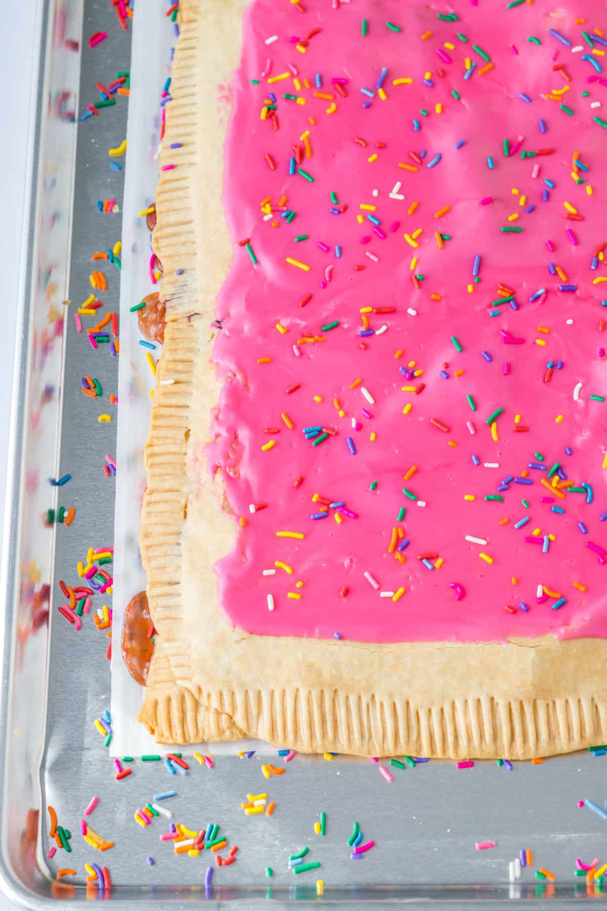 Homemade giant strawberry pop tart made with pie crust, strawberry jam, pink icing, and rainbow sprinkles on a sheet pan.