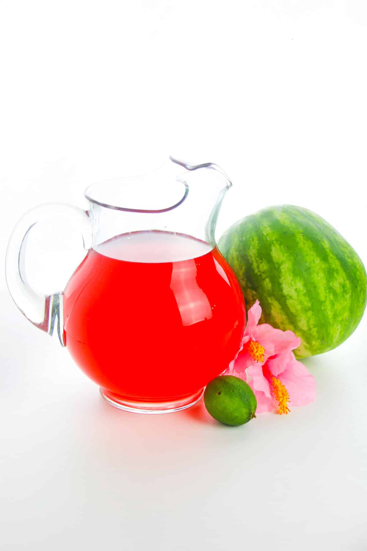 Watermelon drink in large glass pitcher with a small watermelon, a lime, and a pink flower next to it.