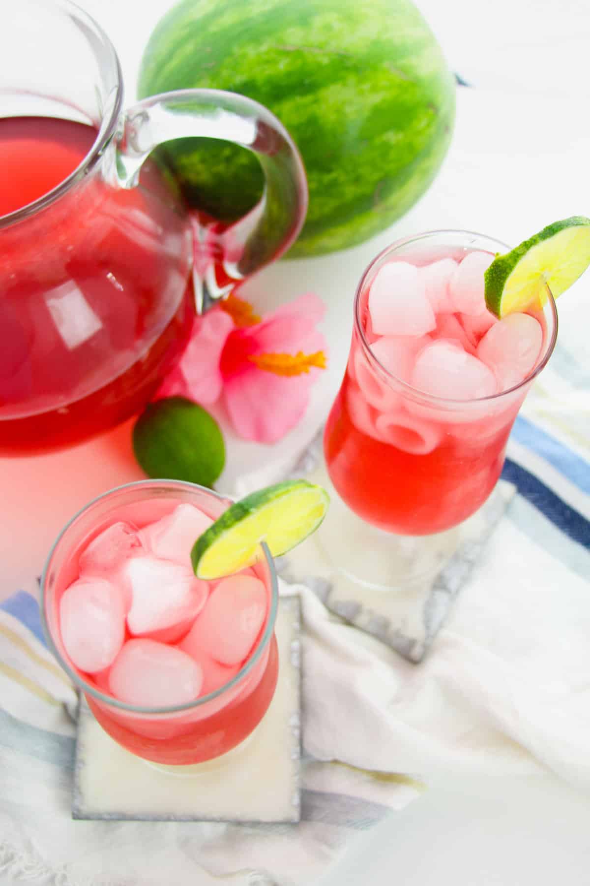 Two watermelon drinks garnished with lime next to watermelon and pitcher.