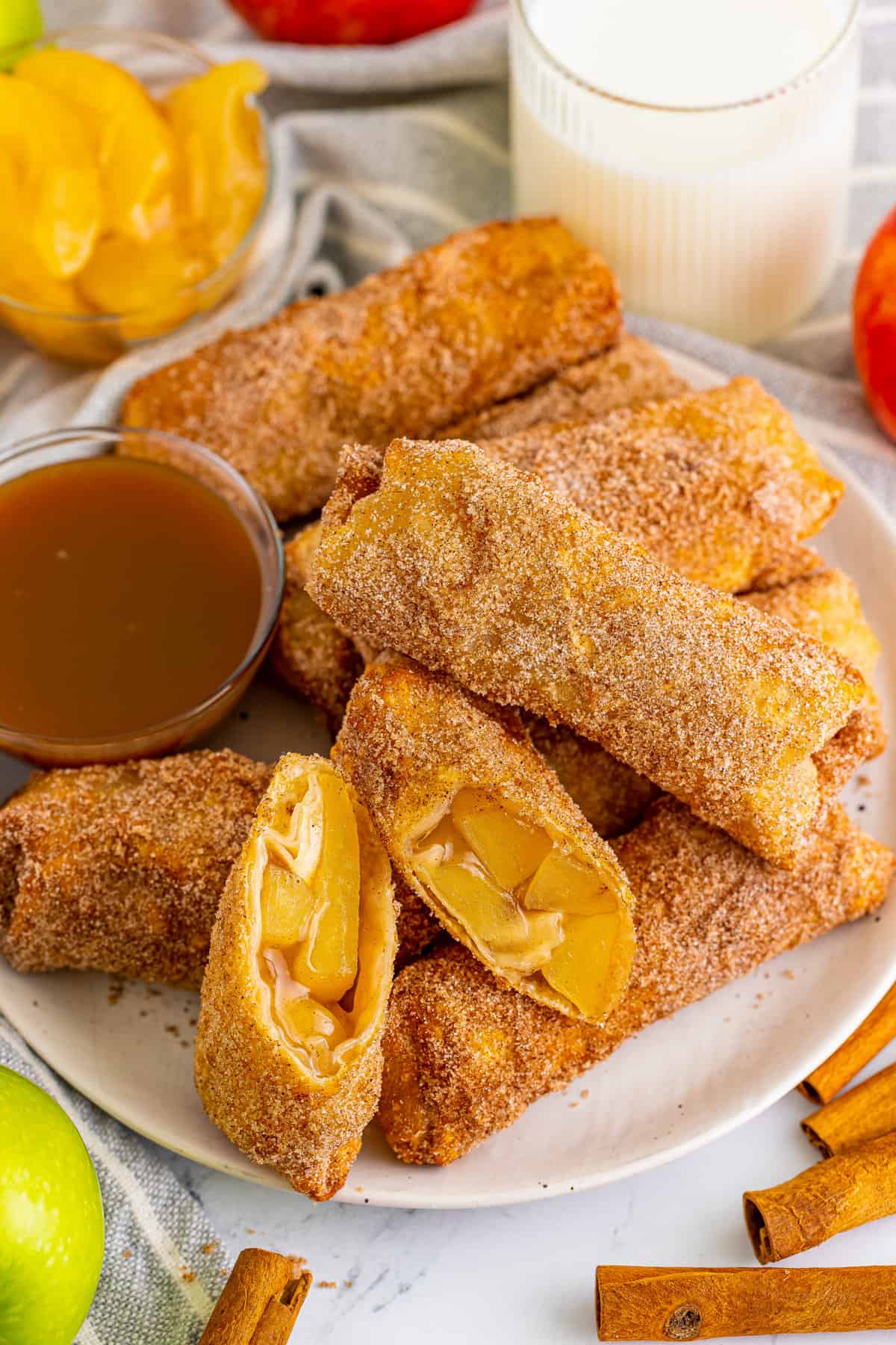 Apple pie egg roll with a bowl of caramel sauce on a plate. One apple egg roll is cut in half to show apple pie filling inside.