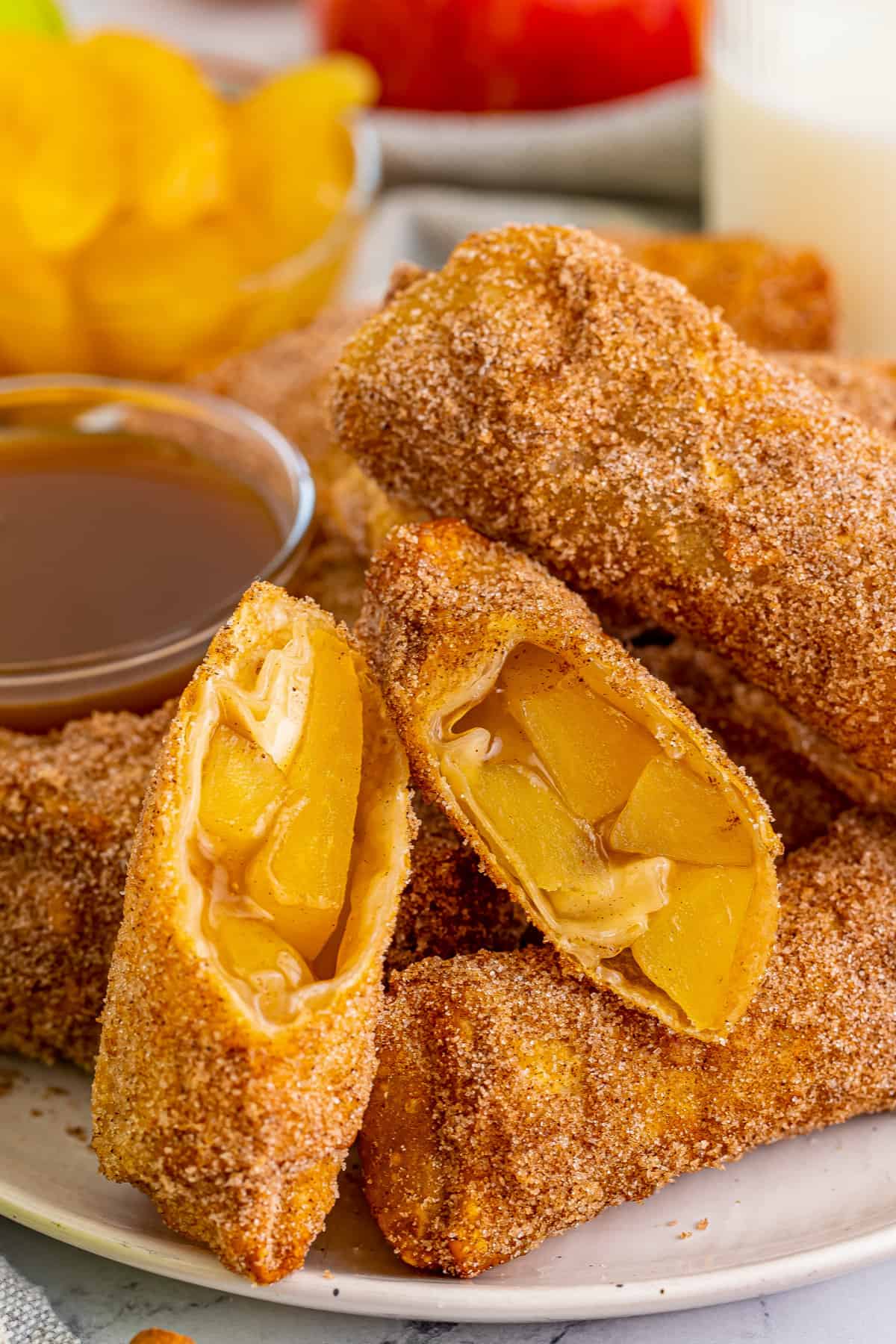Apple pie egg rolls coated in cinnamon sugar and served with caramel sauce for dipping. One egg roll is cut in half to show apple pie filling.