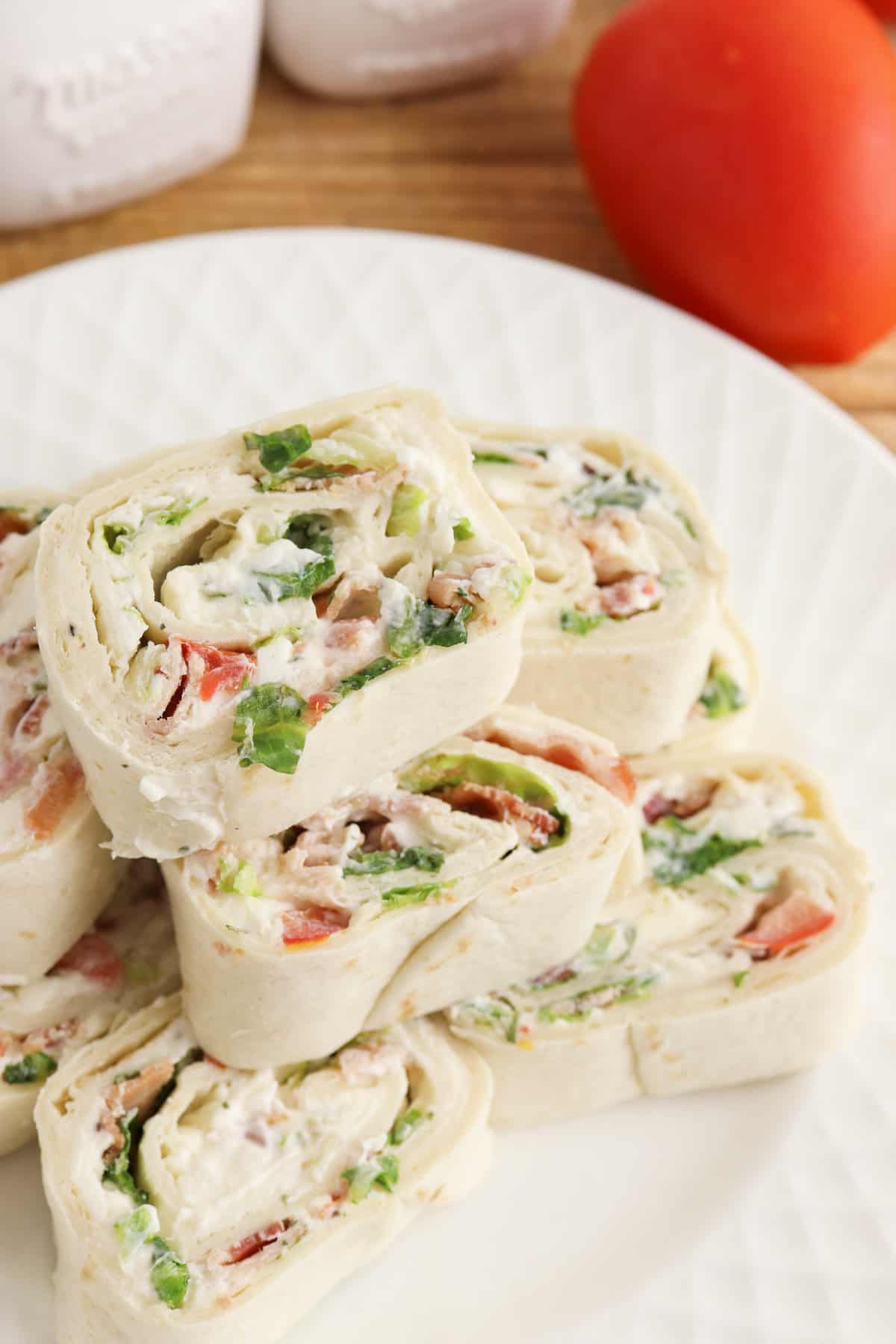 Ranch BLT Pinwheels made with flour tortillas, bacon, lettuce, tomatoes, and ranch dressing stacked on a white plate with a plum tomato next to it.