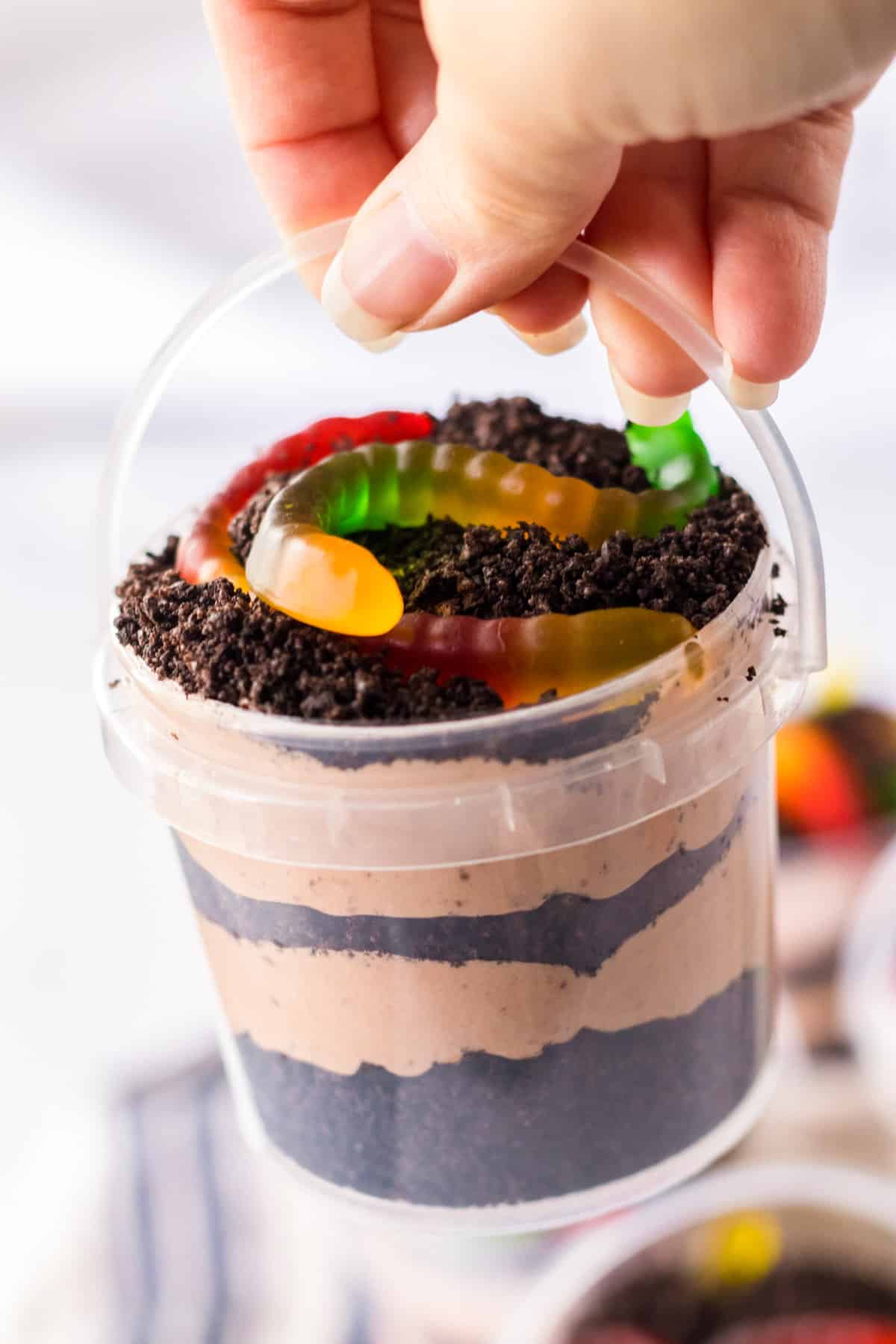 Hand holding oreo dirt pudding with gummy worms in small clear plastic pail.