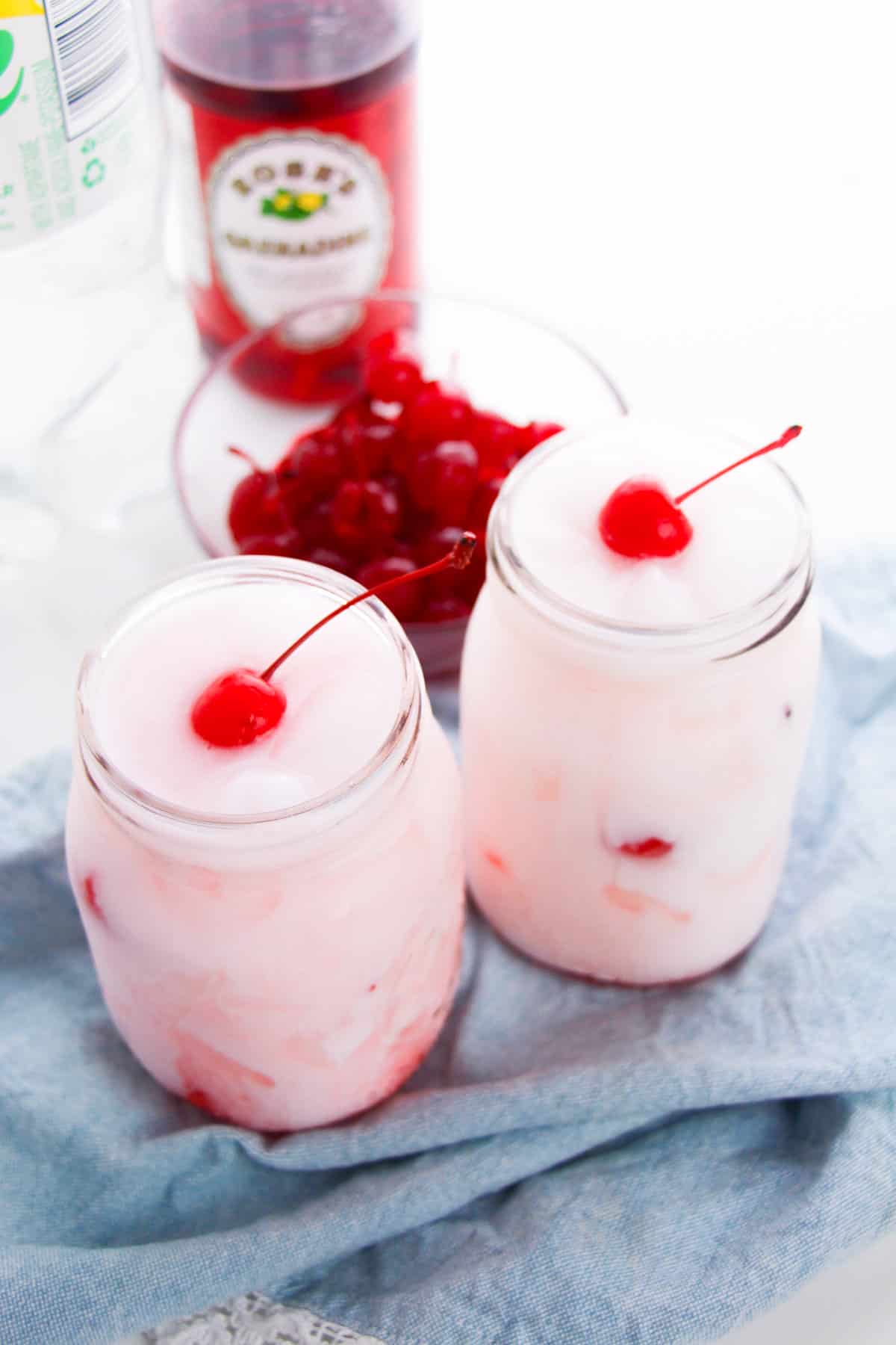 Two Shirley Temple Dirty sodas in mason jars with a bottle of grenadine in the background.