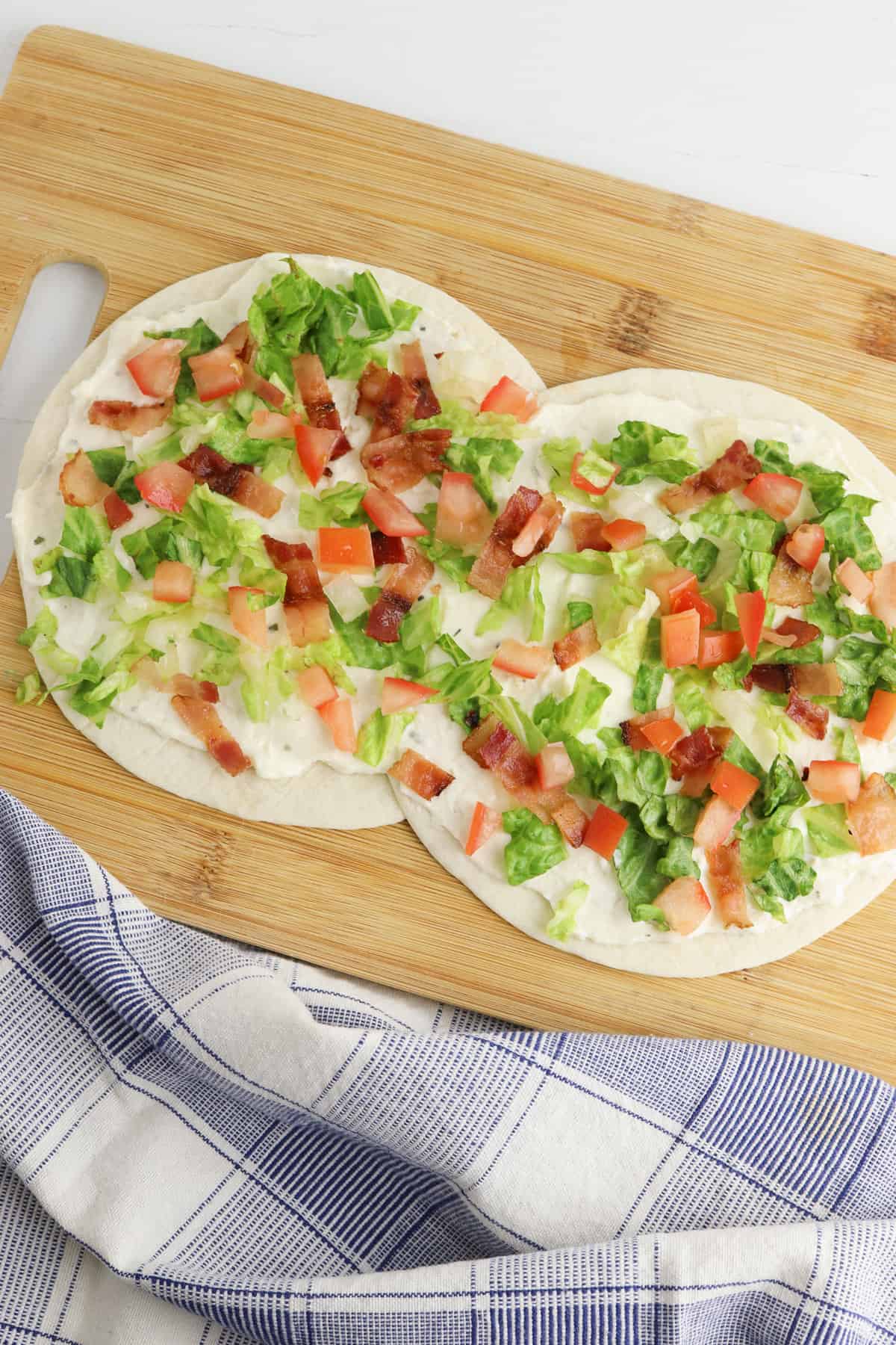 Two flour tortillas layed slightly overlapping on a cutting board. The tortillas are topped with creamy dressing, shredded lettuce, diced tomatoes, and chopped bacon.