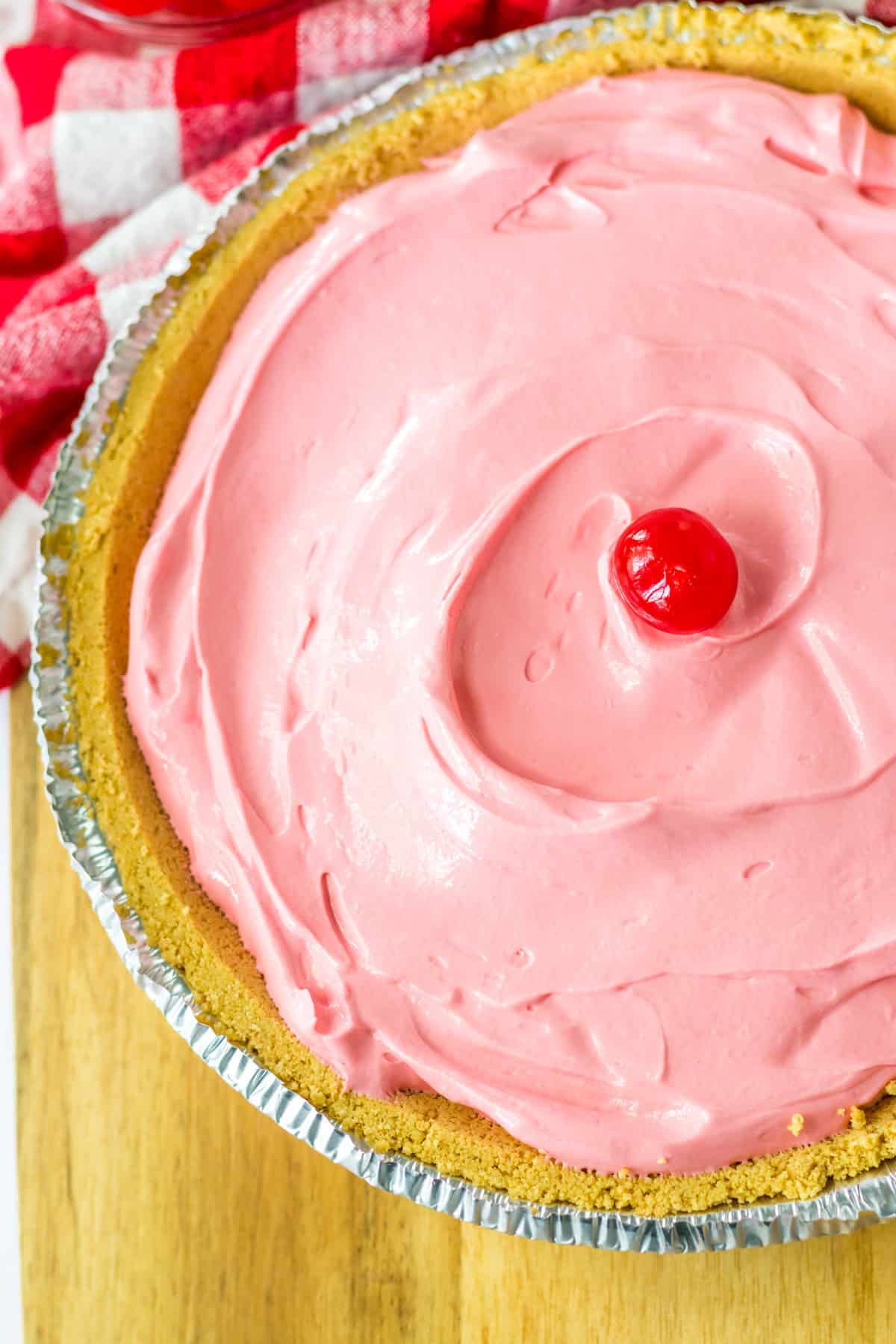 Overhead photo of pink, creamy no bake kool aid pie in graham cracker crust with maraschino cherry in the center.