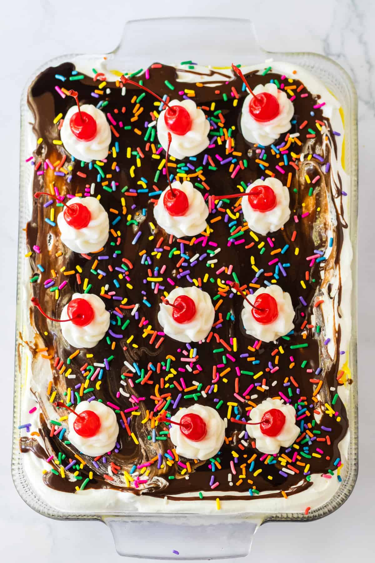Dessert topped with chocolate syrup, rainbow sprinkles, and 12 dollops of whipped cream each topped with maraschino cherries.