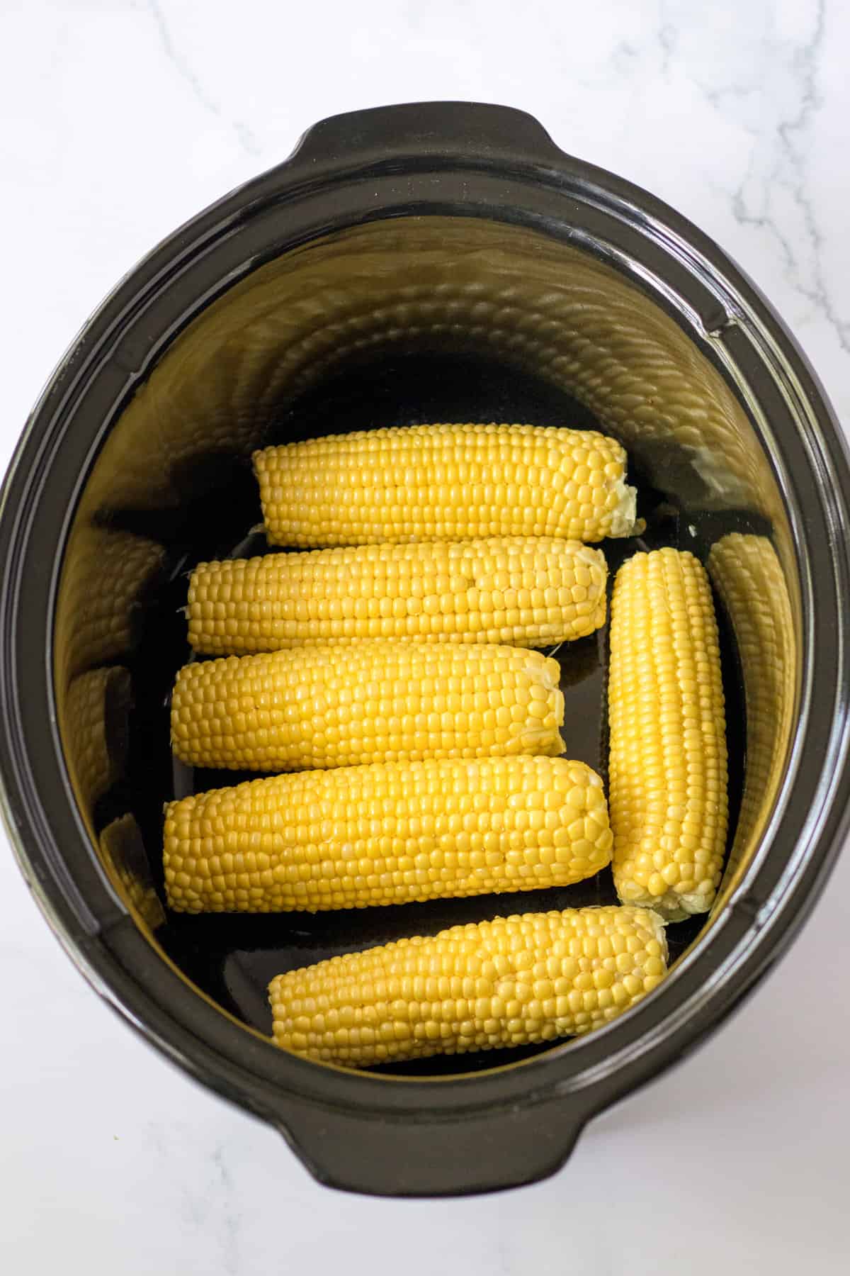 Uncooked corn on the cob in an even layer in the bottom of a slow cooker.