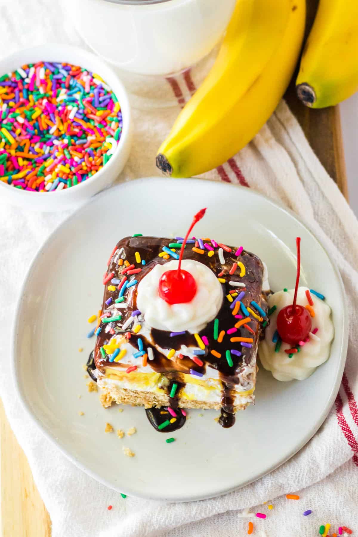 Square piece of no bake banana split cake on white plate with whipped cream and a cherry. Rainbow sprinkles and fresh bananas are next to the plate.