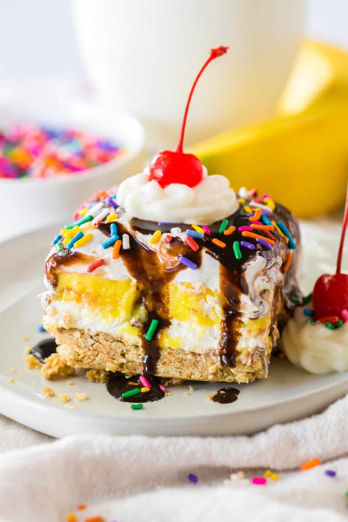 No bake banana split dessert with a graham cracker crust; layers of sweetened cream cheese, banana pudding, and Cool Whip; and topped with chocolate syrup, whipped cream, and a cherry.