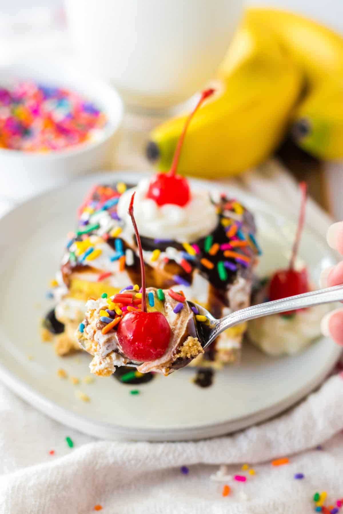 Spoonful of banana pudding lush dessert with slice, bananas, and sprinkles in background.