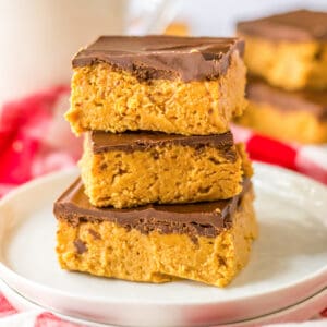 Three no-bake chocolate peanut butter bars stacked on a plate with milk and more bars behind them.