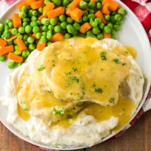 Pork chops smothered in gravy served over mashed potatoes and with a side of peas and carrots.