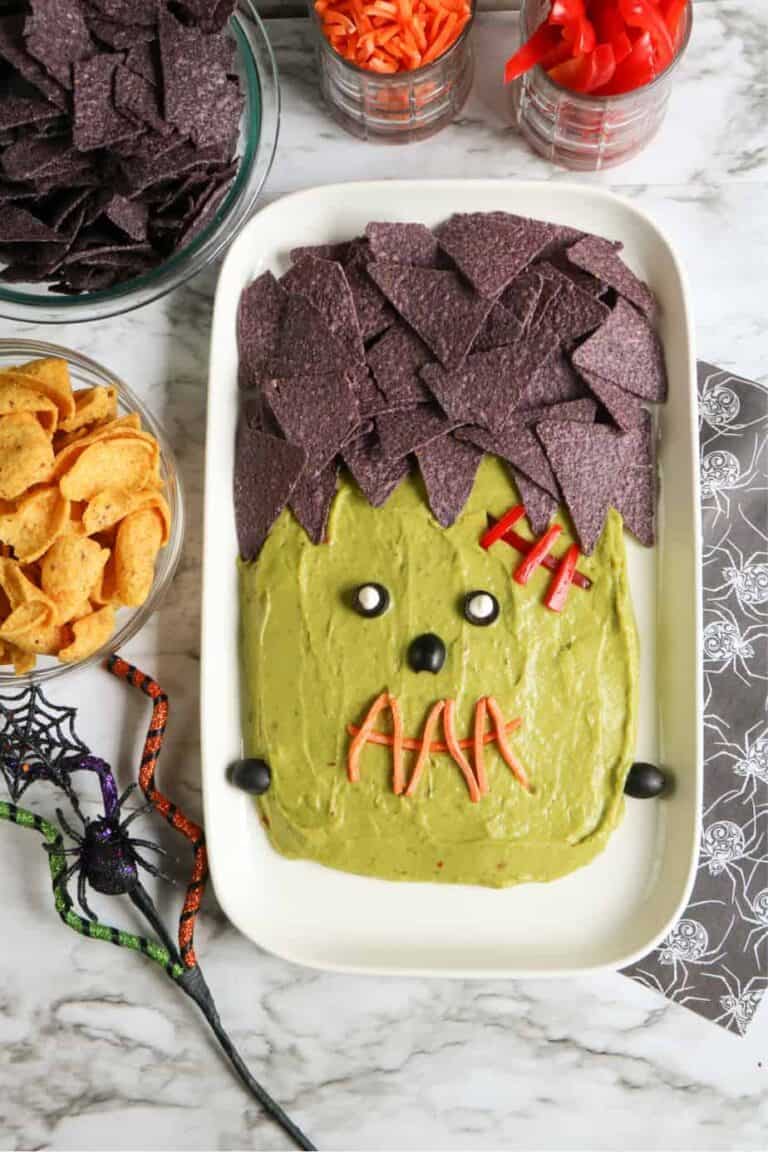 Frankenstein guacamole decorated with black tortilla chips, carrot shreds, olives, and sliced peppers.