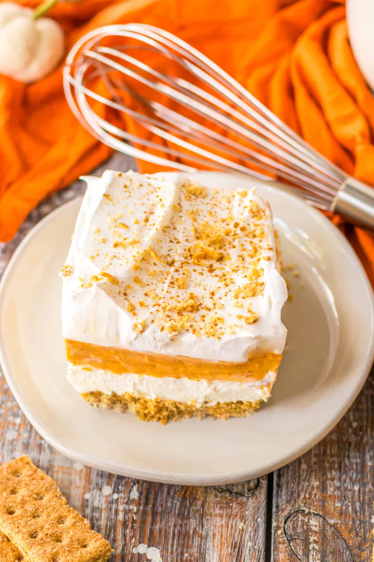 Square piece of layered pumpkin dessert on white plate with wire whisk and orange linen.