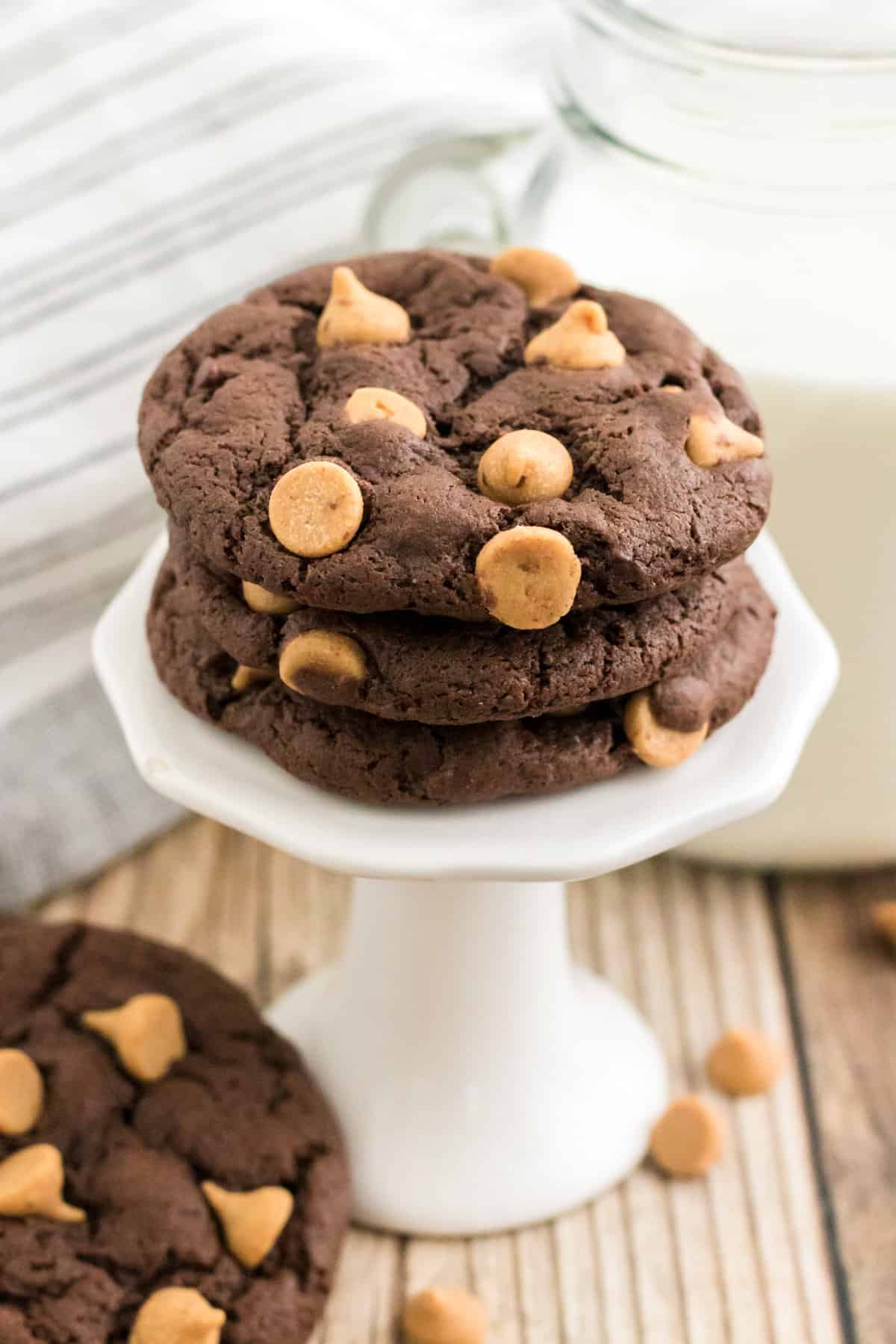 Three peanut butter chip chocolate cookies stacked on a small white stand with a glass of milk behind them.