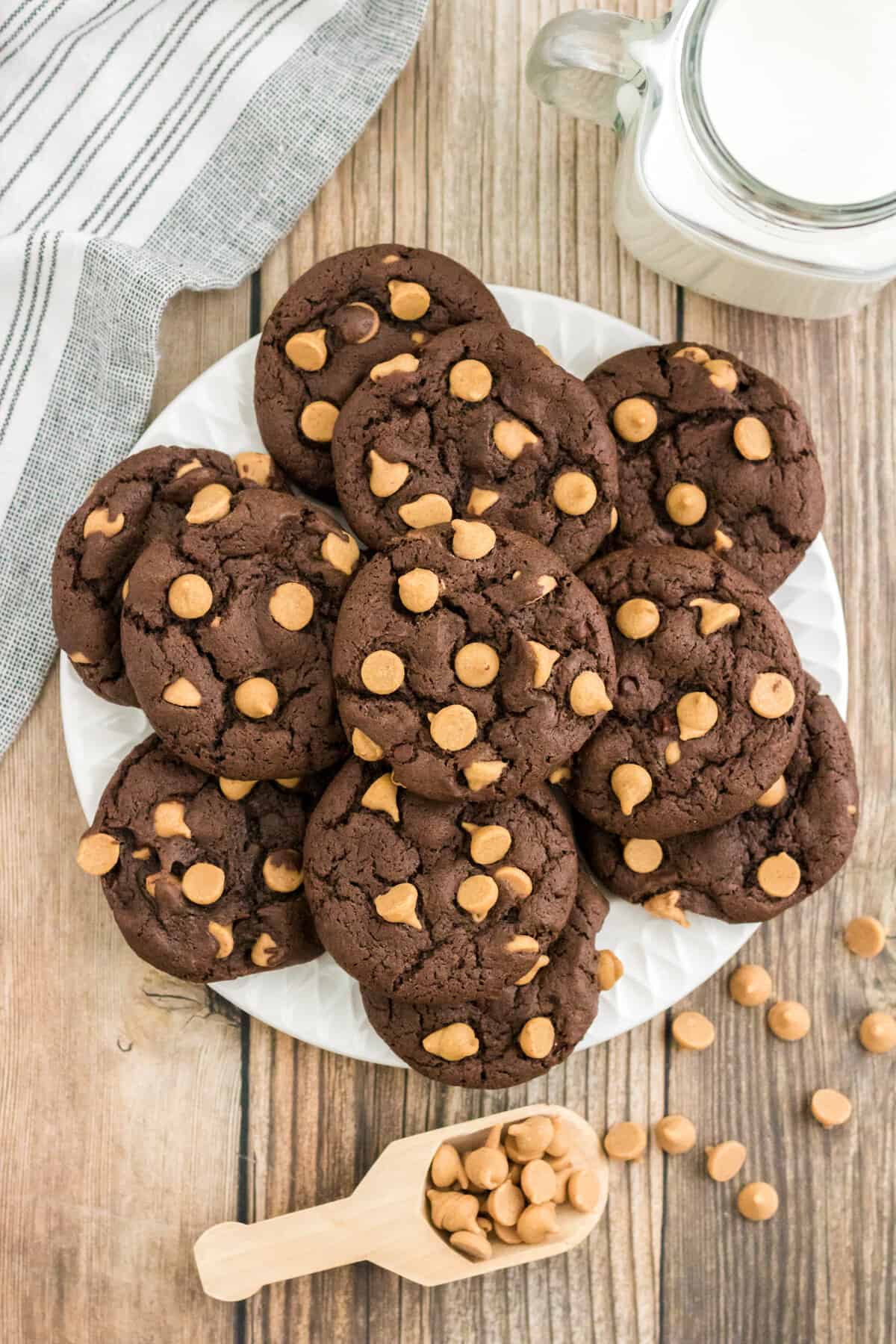 Chocolate peanut butter chip cookies on a white plate with peanut butter morsels and a glass of milk beside them.