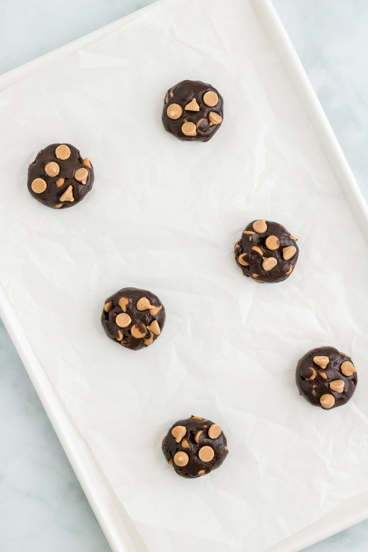 Six balls of chocolate cookie dough with peanut butter chips pressed into the tops spaced apart on a parchment-lined baking sheet.