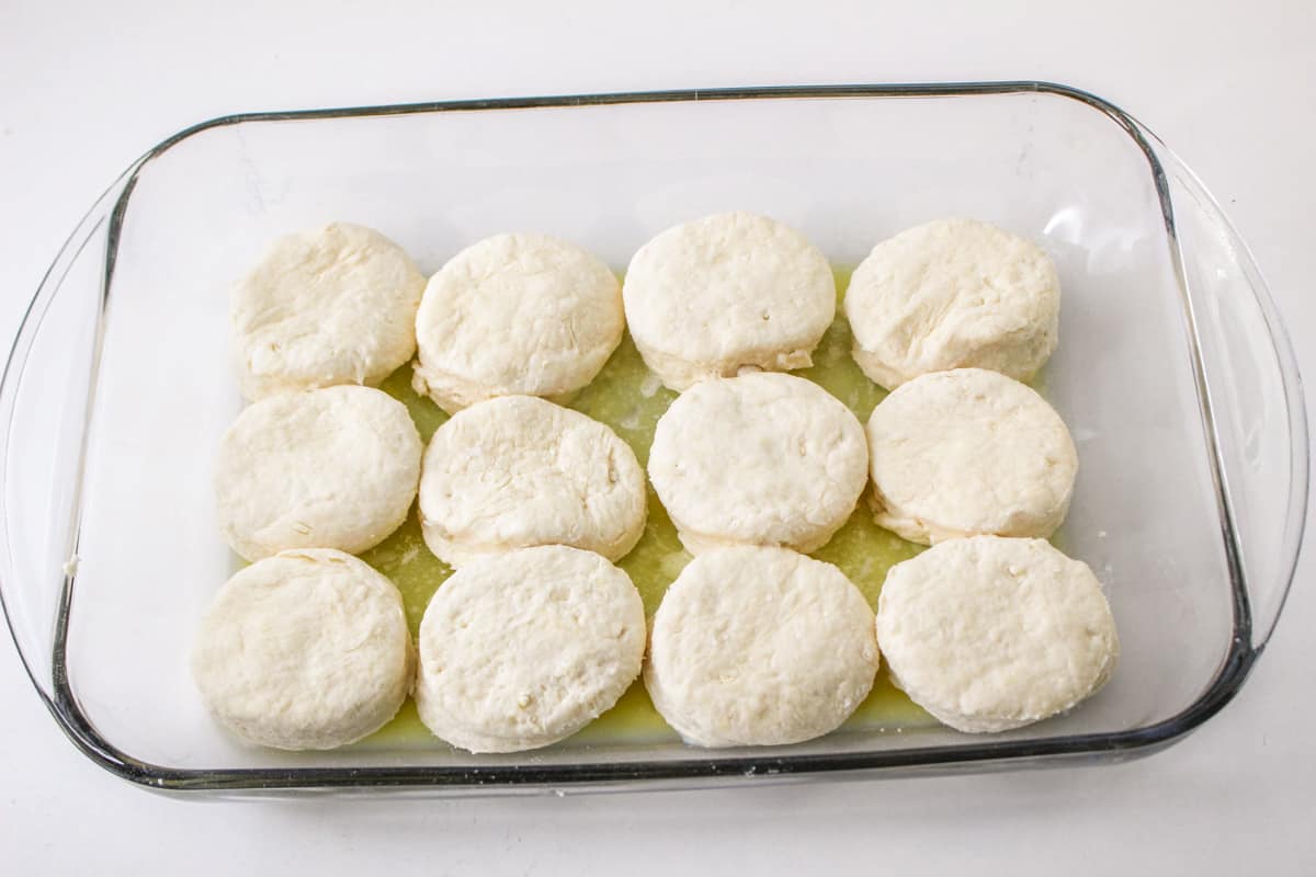 Raw biscuits on top of butter in baking dish.