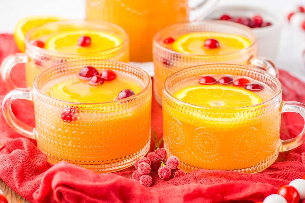 Four glass mugs of non-alcoholic Christmas punch garnished with orange slices and cranberries.