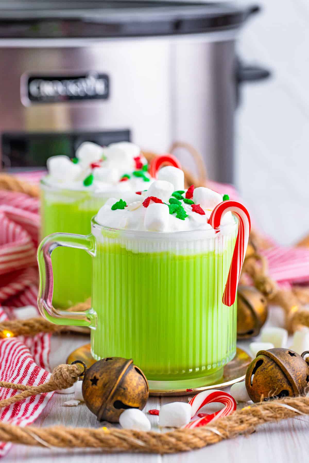 Slow cooker grinch hot chocolate in two glass mugs with whipped cream, mini marshmallows, christmas sprinkles, and mini candy canes. A crockpot is in the background.