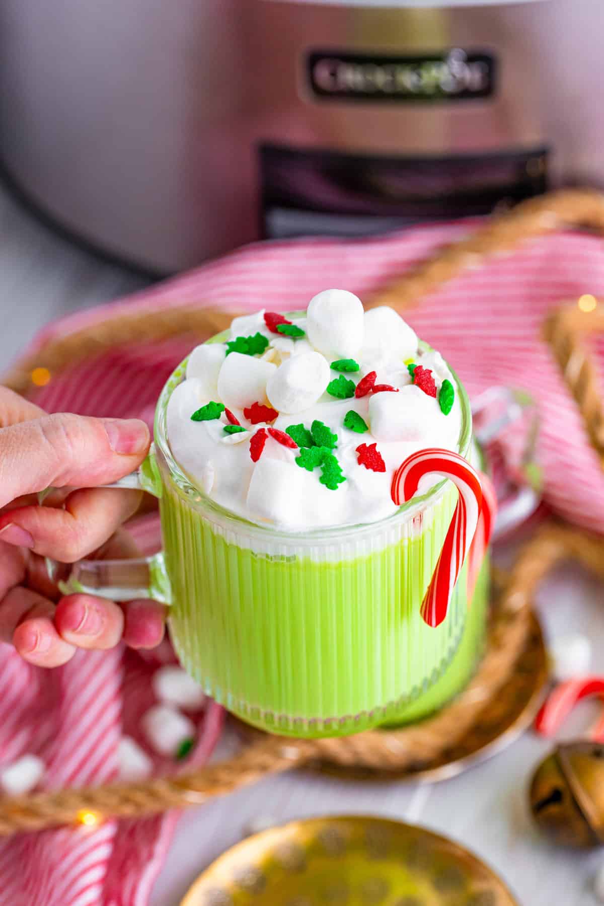 Hand holidaying mug of Grinch hot chocolate with a slow cooker in the background.