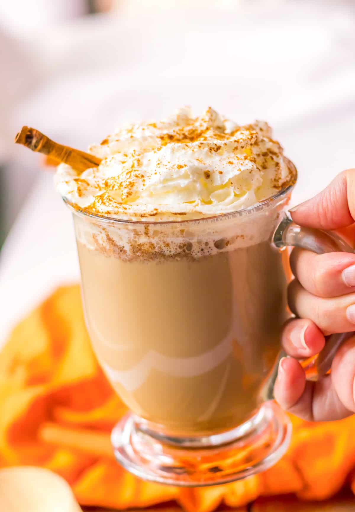 Pumpkin spice latter in a glass mug topped with a big pile of whipped cream, a sprinkle of cinnamon, and a cinnamon stick.
