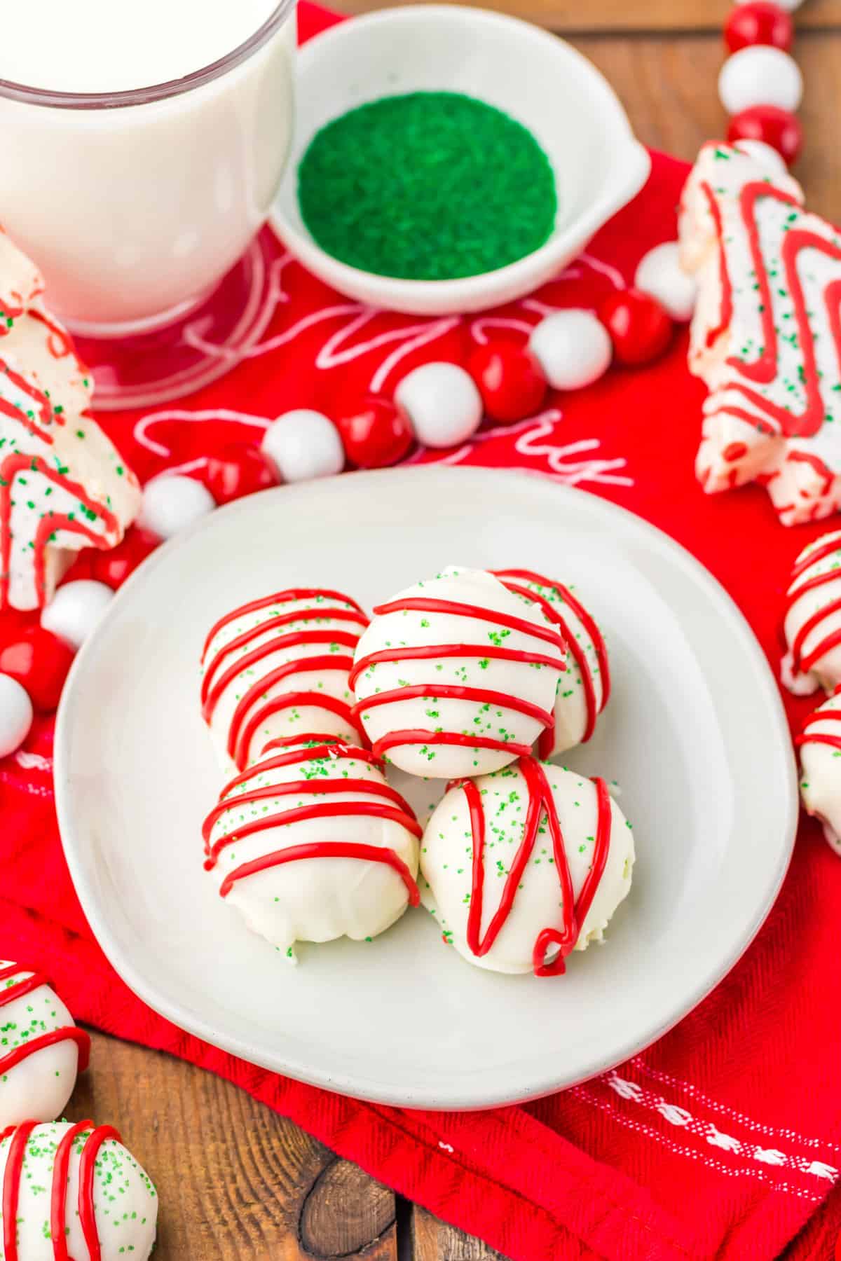 Little Debbie Christmas Tree Cake Balls on a white plate with a glass of milk, green sugar sprinkles, and Little Debbie Christmas Tree Snack Cakes in the background.