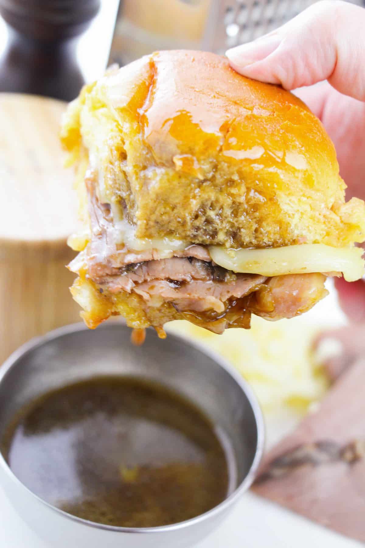 Hand holding French dip roast beef slider and dipping it in au jus sauce.
