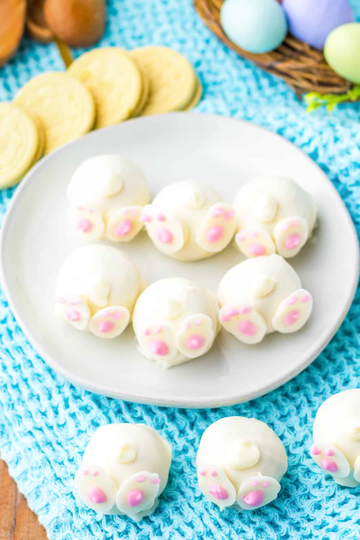 Overhead view of bunny truffles coated with white chocolate and decorated with candy feet and tails. Lemon cookies and easter decorations are surrounding them.