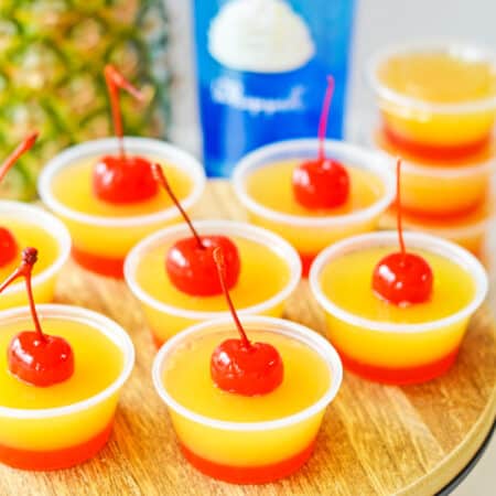 Pineapple upside down cake jello shots with two distinct layers: a red cherry bottom layer and yellow pineapple top layer, and a cherry on top.