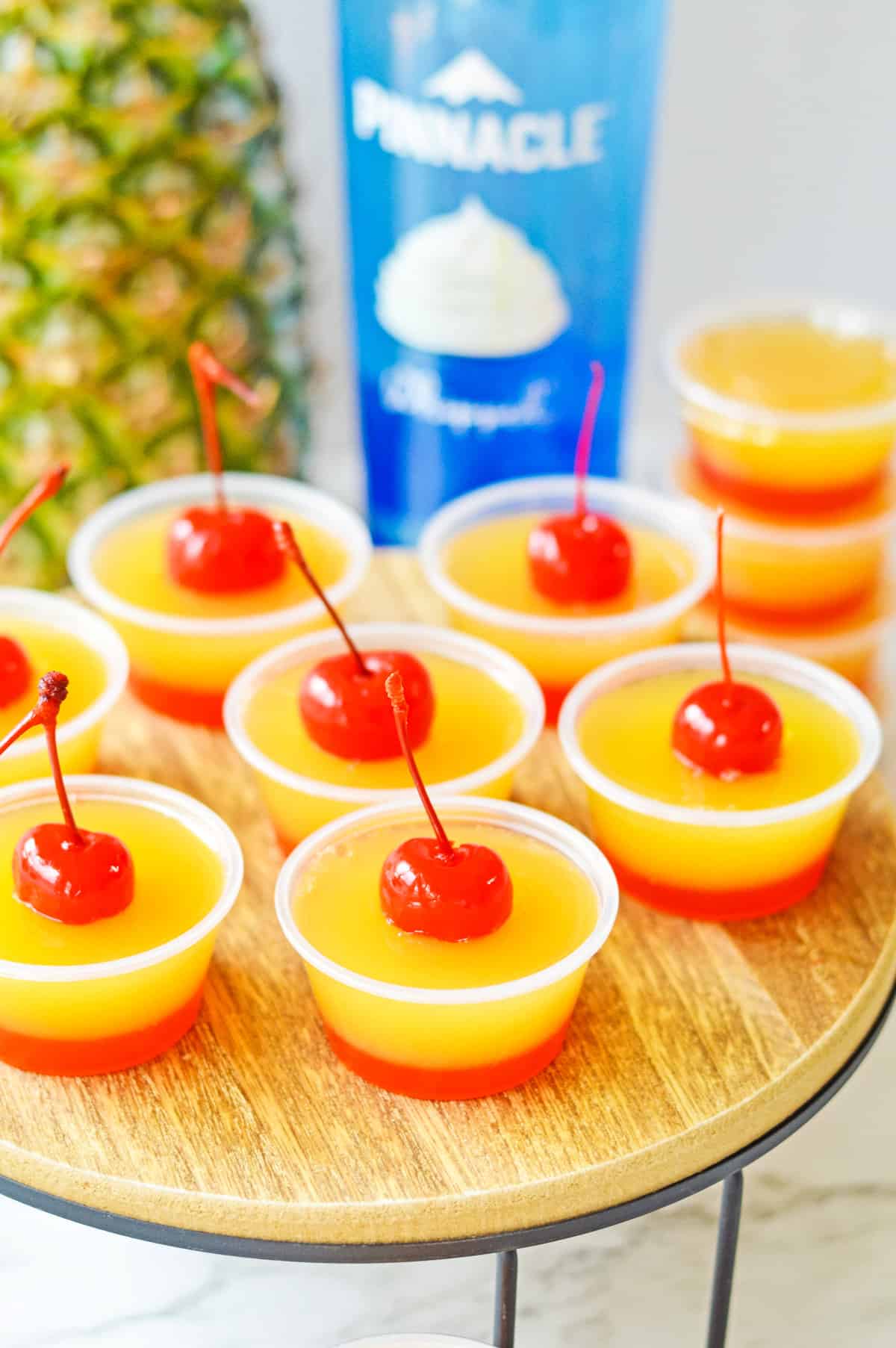 Pineapple upsid down cake jello shots with two distinct layers: a red cherry bottom layer and yellow pineapple top layer, and a cherry on top.