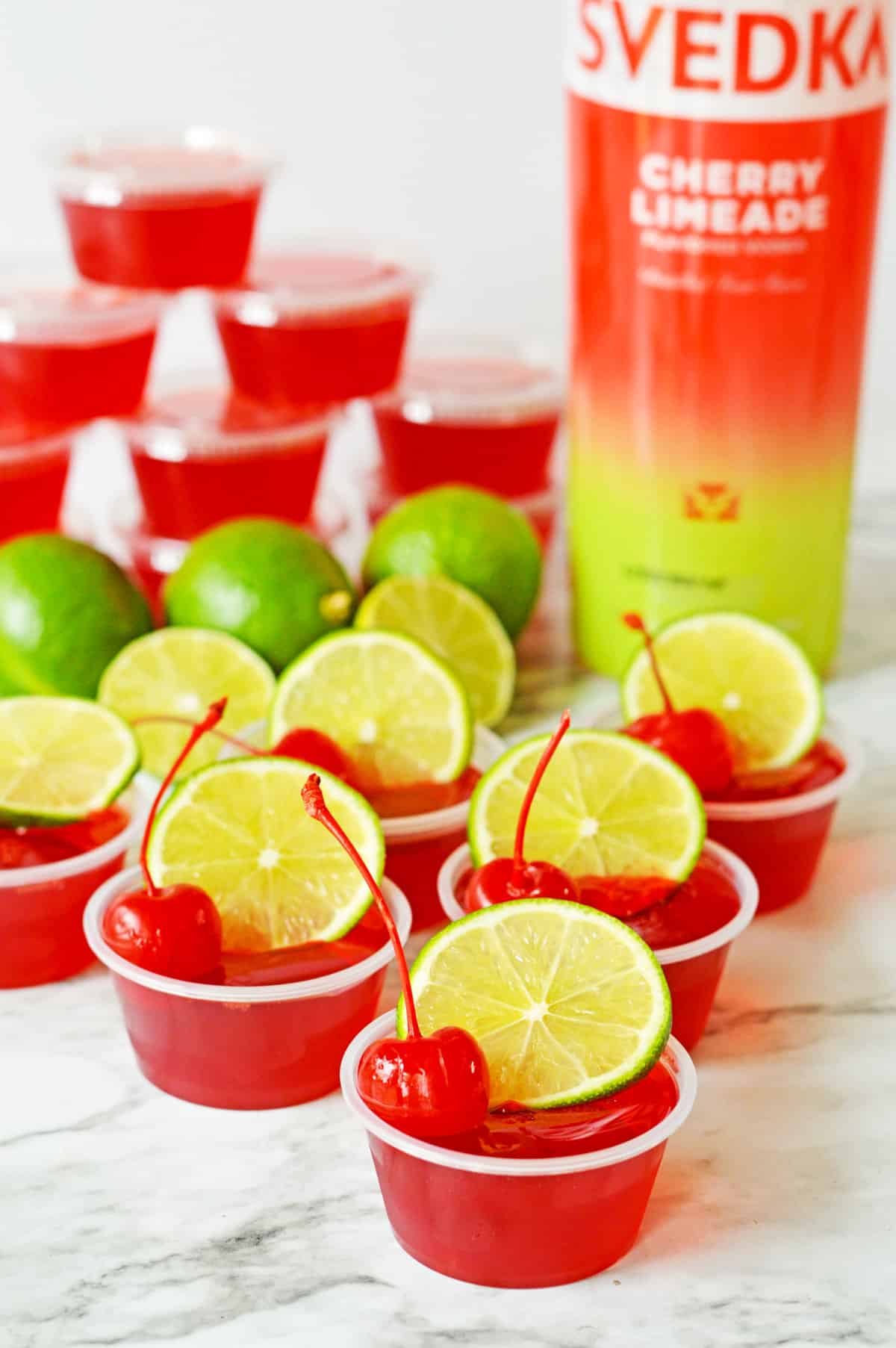 Cherry Limeade Jello Shots made with red jello and garnished with lime and maraschino cherries. Fresh limes, shots covered with lids, and a bottle of SVEDKA cherry limeade vodka are in the background.