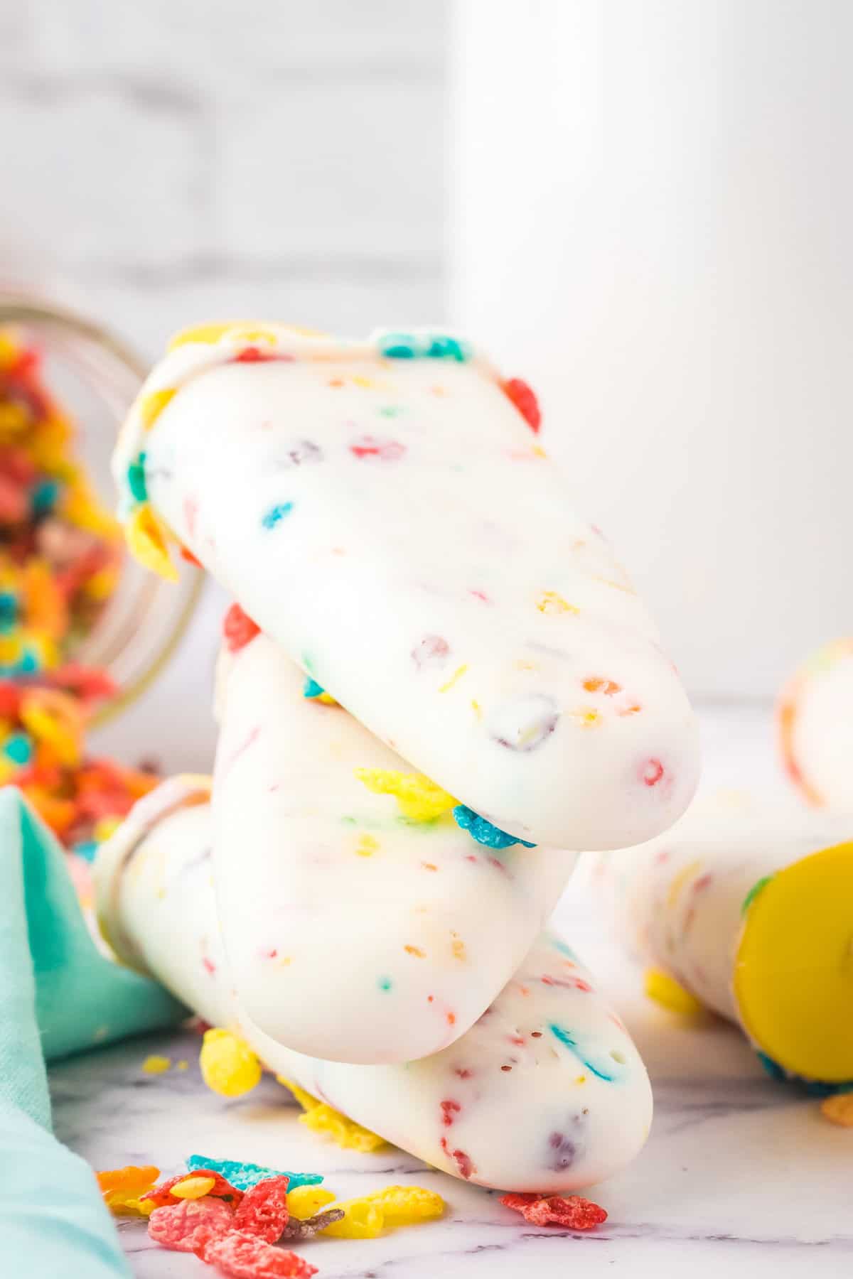 Rainbow cereal and yogurt breakfast popsicles stacked on top of one another in front of a large glass of milk.