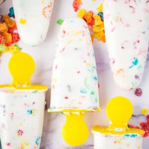 Homemade fruity Pebbles cereal Greek yogurt popsicles with yellow handles. The colorful cereal can be seen throughout the white ice pops.