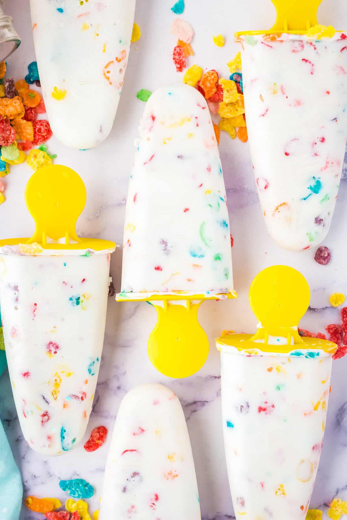 Homemade fruity Pebbles cereal Greek yogurt popsicles with yellow handles. The colorful cereal can be seen throughout the white ice pops.