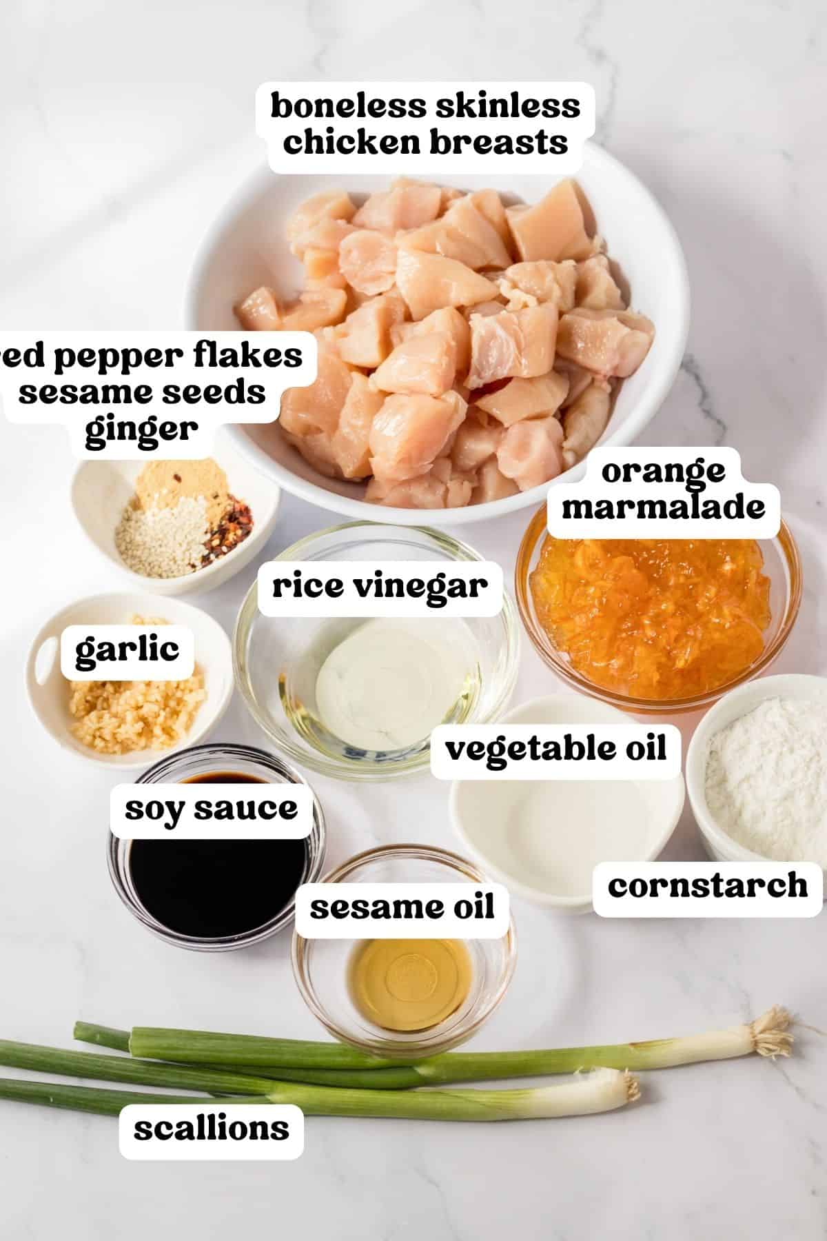 Ingredients in bowls on countertop: boneless skinless chicken breasts cubed, orange marmalade, rice vinegar, minced garlic, sesame oil, soy sauce, cornstarch, vegetable oil, red pepper flakes, ground ginger, sesame seeds, and fresh scallions.