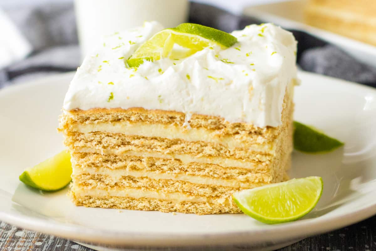Square slice of key lime icebox cake with layers of key lime custard and graham crackers topped with whipped cream and garnished with key lime slice.