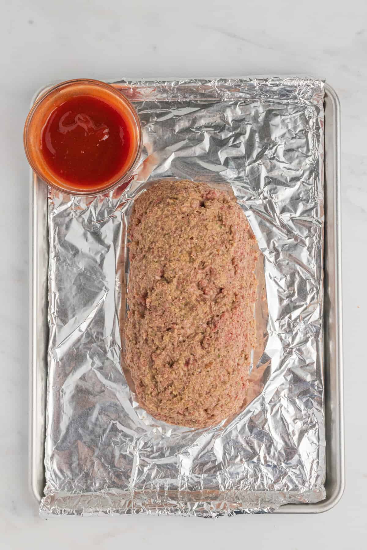 Meatloaf formed into loaf shape on foil lined baking sheet with a bowl of glaze beside it.