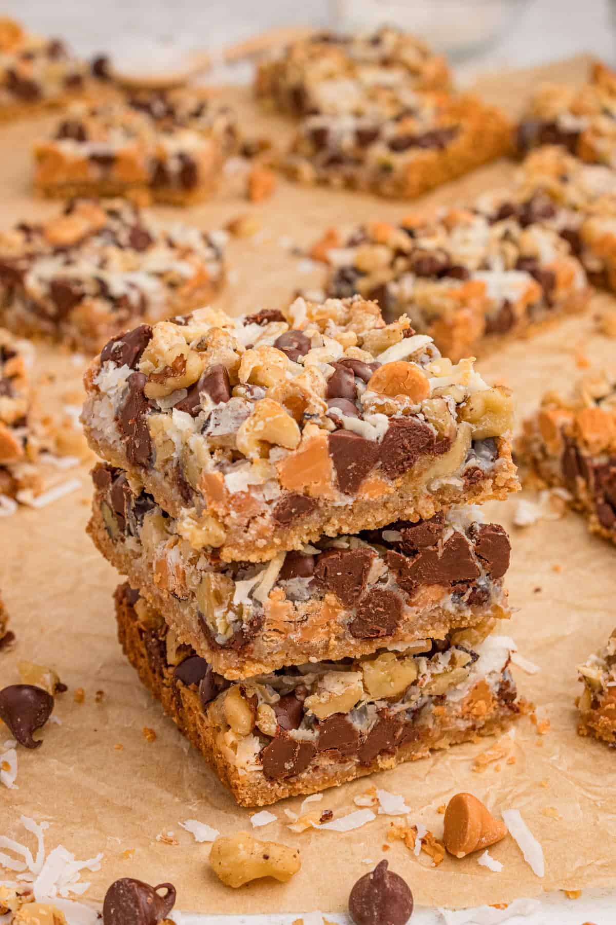 7 Layer bars with chocolate chips, butterscotch chips, shredded coconut, and chopped walnuts over a graham cracker crust. Three of the cookie bars are stacked on top of each other with additional bars in the background.