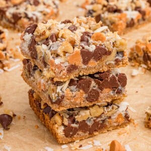 7 Layer bars with chocolate chips, butterscotch chips, shredded coconut, and chopped walnuts over a graham cracker crust. Three of the cookie bars are stacked on top of each other with additional bars in the background.