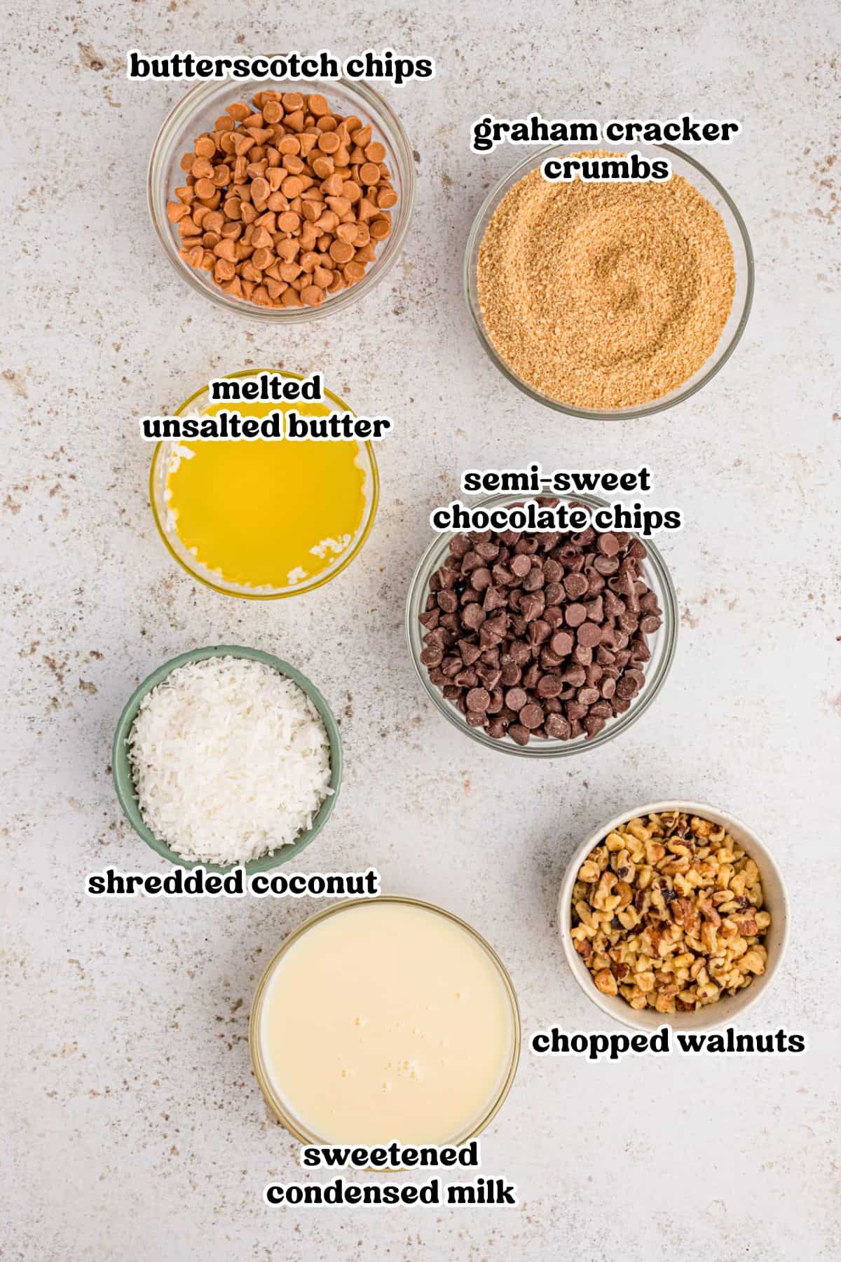 Butterscotch chips, chocolate chips, graham cracker crumbs, shredded coconut, chopped walnuts, sweetened condensed milk, and melted butter in bowls.