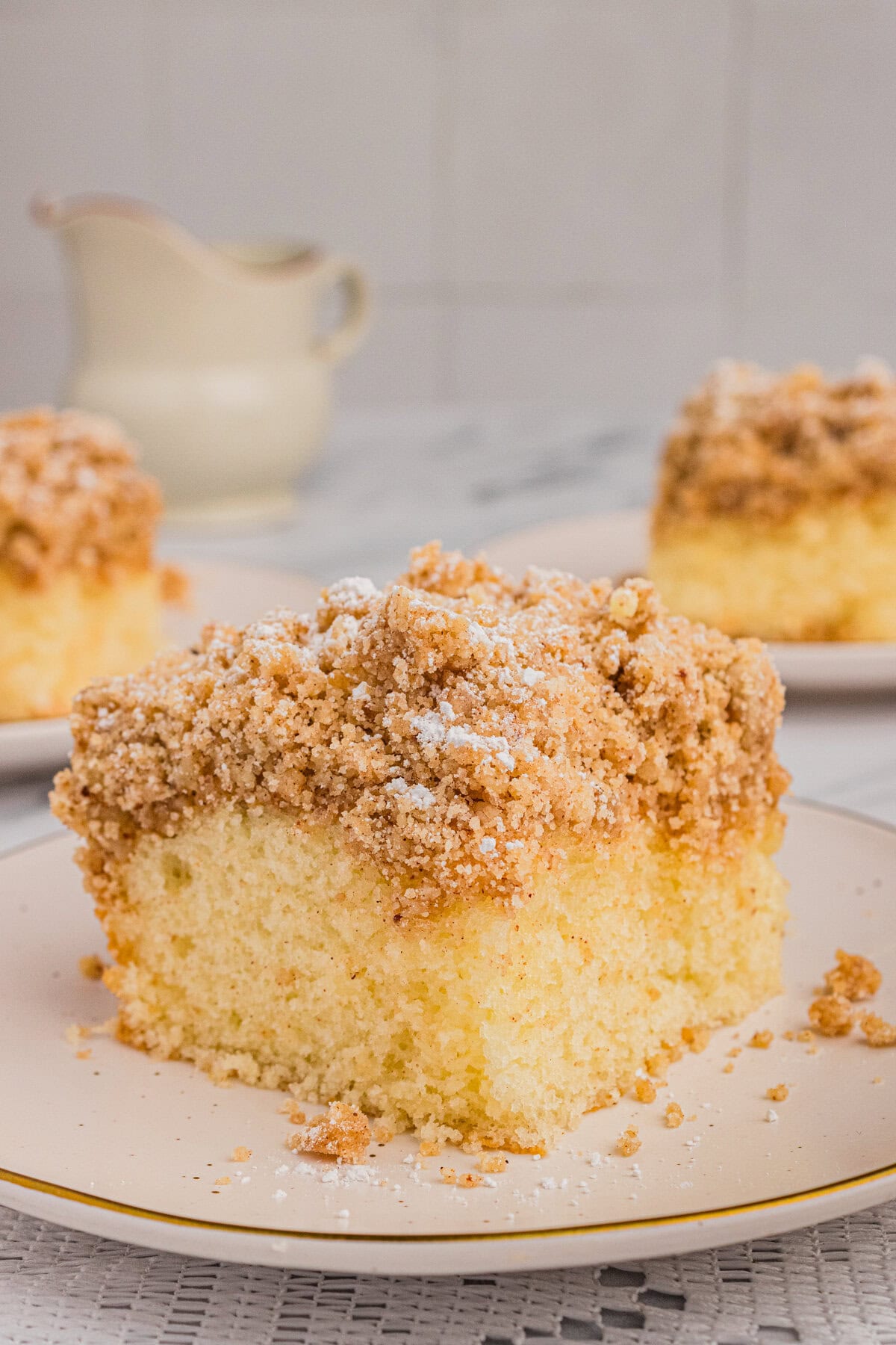 Slice of crumb cake with corner facing the camera and a teapot in the background.