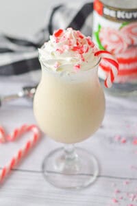 A creamy boozy peppermint milkshake in a glass, topped with whipped cream, crushed peppermint candies, and a candy cane garnish and a festive background.