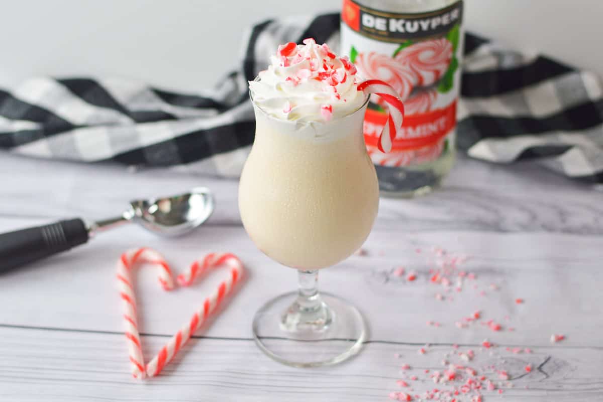 A creamy peppermint schnapps milkshake in a glass, topped with whipped cream, crushed candy canes, and a candy cane garnish, with a bottle of peppermint schnapps in the background.