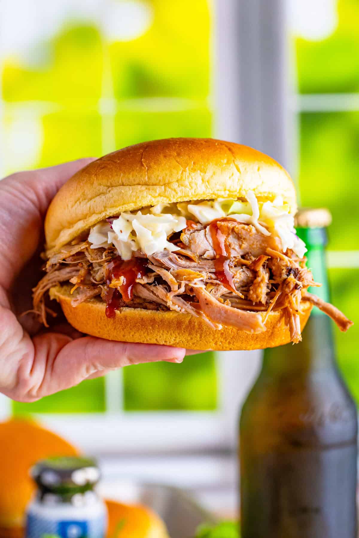 A hand holding a pulled pork sandwich made with a soft bun, topped with shredded pork, creamy coleslaw, and a drizzle of BBQ sauce.