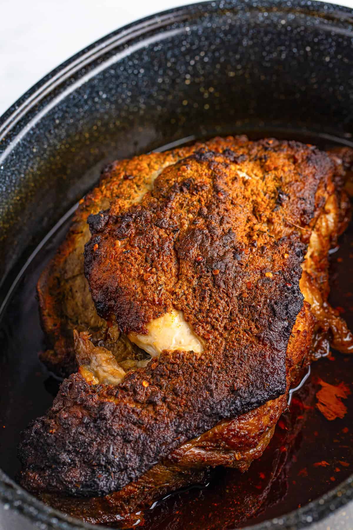 A fully cooked pork shoulder in a roasting pan, featuring a crispy, caramelized crust and juices pooling at the bottom of the pan.