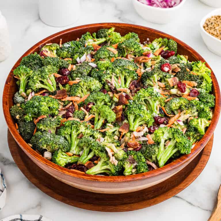 A large wooden bowl filled with broccoli crunch salad, made with fresh broccoli, bacon bits, dried cranberries, shredded carrots, sunflower seeds, and chopped red onions, all lightly coated in a creamy dressing.