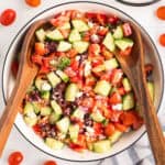 A large bowl of colorful Greek salad, filled with chopped cucumbers, cherry tomatoes, red bell peppers, kalamata olives, red onion, crumbled feta, and herbs. Two wooden salad servers rest on the salad.